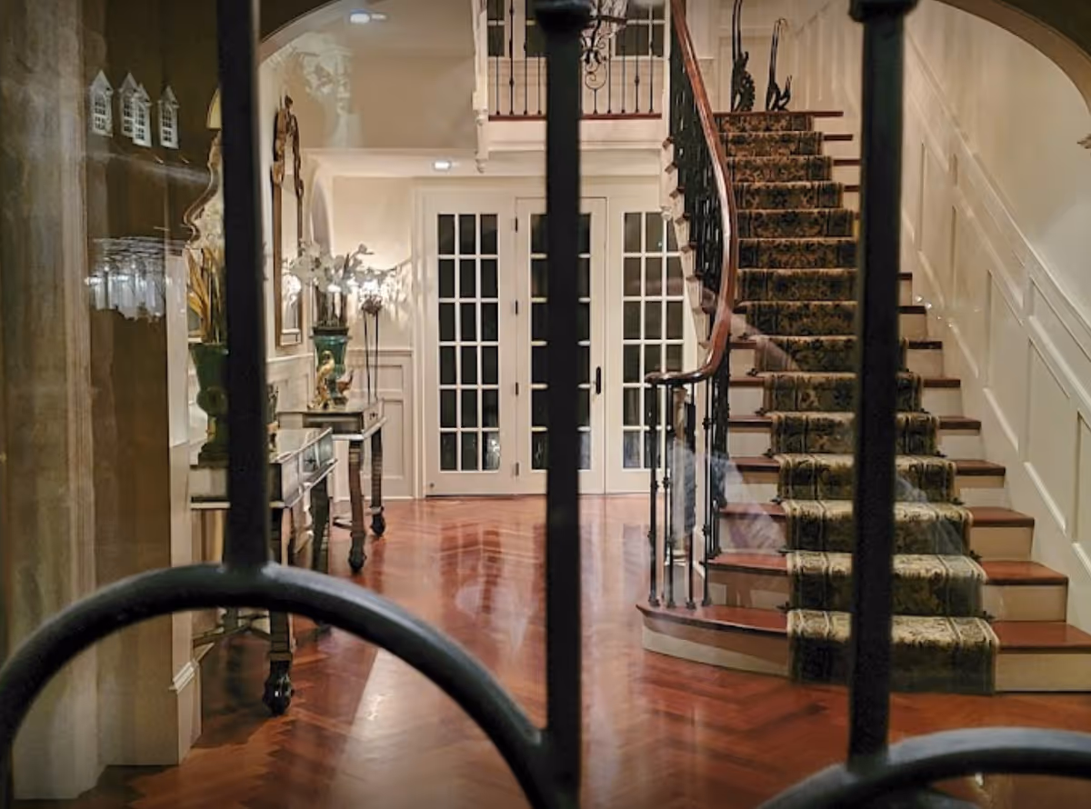 View through a decorative wrought iron gate into an elegant interior hallway featuring polished wooden floors, a carpeted staircase with ornate railing, a console table with decorative items and flowers, and double glass-paneled doors at the far end.