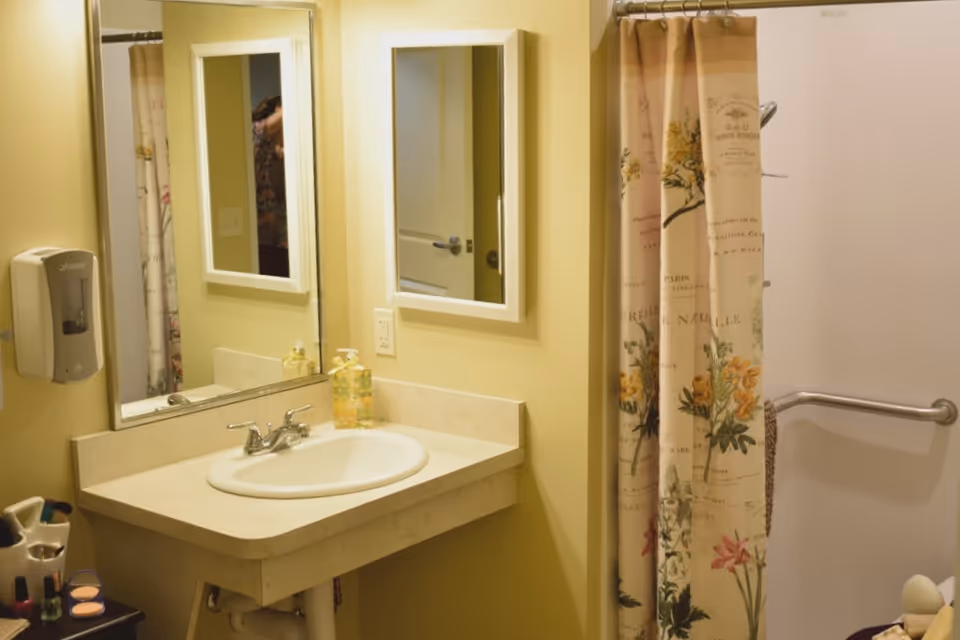 A bathroom with a beige countertop and white sink, a wall-mounted soap dispenser, two mirrors above the sink, and a shower area with a floral patterned shower curtain and a grab bar on the wall.