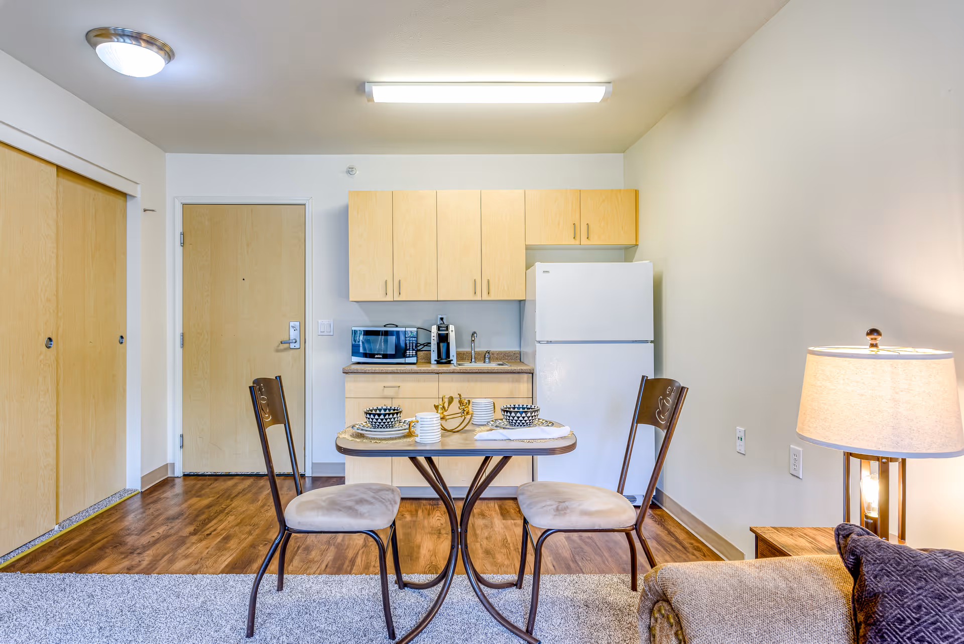A small kitchen area in a senior living facility with light wood cabinets, a white refrigerator, a microwave, and a coffee maker. In front of the kitchen is a small dining table set for two with bowls, plates, and cups. To the right, there is a beige couch with a dark pillow and a lit table lamp on a wooden side table. The floor is a combination of carpet and wood, and there is a closed wooden door and closet with sliding doors on the left.