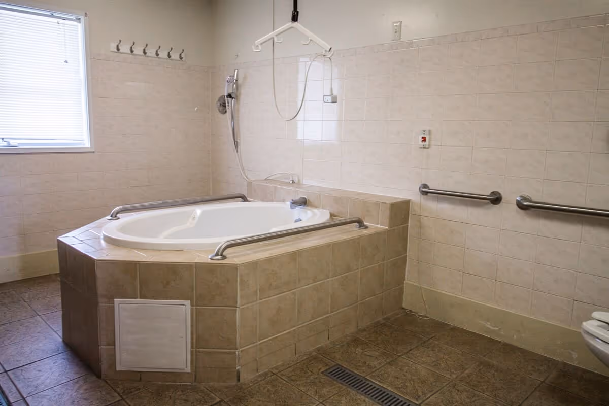 A spacious bathroom with a large, tiled, built-in bathtub equipped with stainless steel grab bars. The walls are tiled halfway up with beige tiles, and there are additional grab bars mounted on the wall. A window with blinds is on the left side, and a showerhead is mounted above the bathtub. The floor is tiled with darker tiles, and there is a floor drain visible.