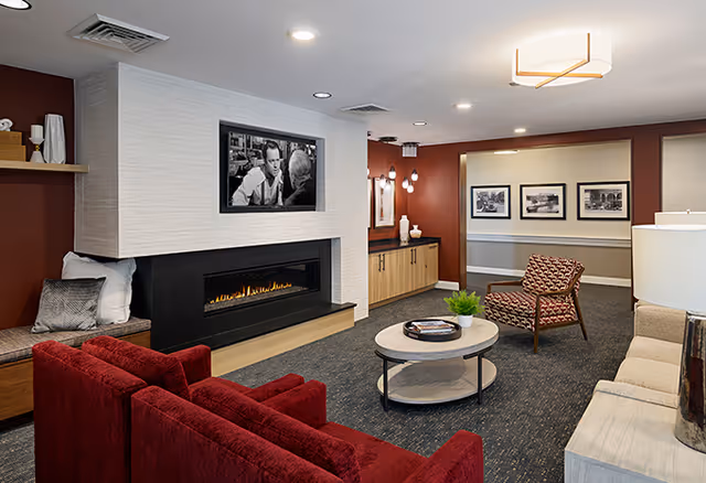 A cozy senior living facility lounge area featuring a modern fireplace with a black and white photo above it, a red sofa, a patterned armchair, a round coffee table with a small plant and books, and a beige sofa with a lamp on a side table. The walls are painted in warm tones with framed black and white photographs and soft lighting fixtures.