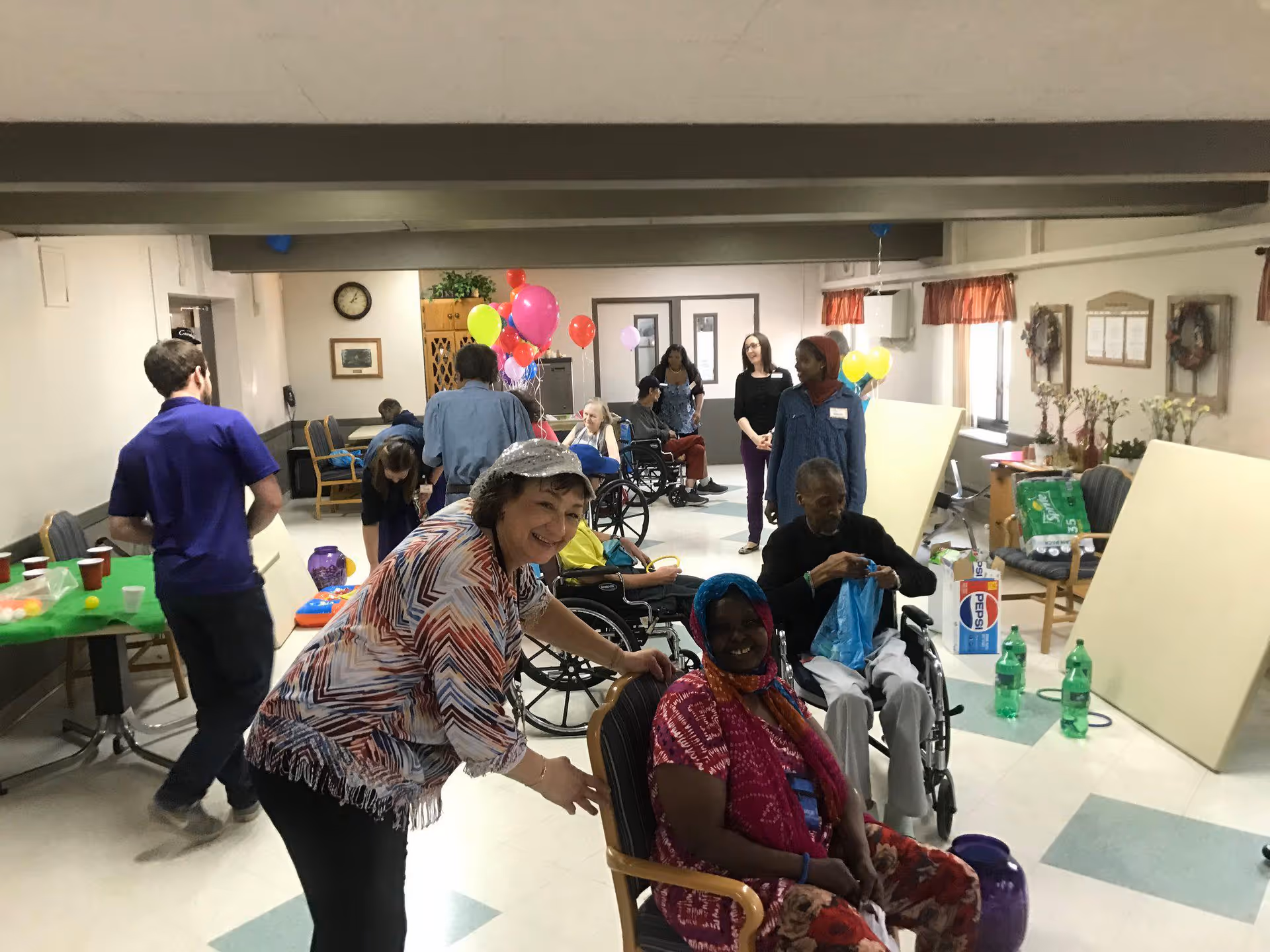 A group of elderly people and caregivers gathered in a common room decorated with colorful balloons. Some individuals are seated in wheelchairs while others stand or sit on chairs. The room has tables with cups and snacks, and the atmosphere appears lively and social.