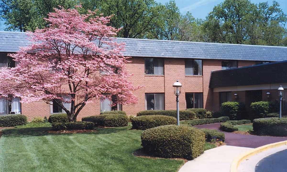 Exterior view of a two-story brick building with several windows, surrounded by neatly trimmed bushes and a well-maintained lawn. A large tree with pink blossoms stands prominently in front of the building. There are lamp posts along a paved walkway leading to the entrance.
