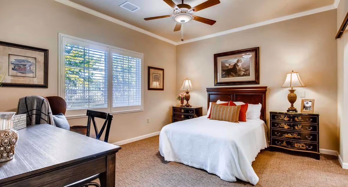 A cozy bedroom with a neatly made bed featuring white bedding and decorative pillows. The room has beige walls, a ceiling fan with a light, two bedside tables with lamps, framed artwork on the walls, a window with white shutters, and a wooden desk with a chair in the foreground.