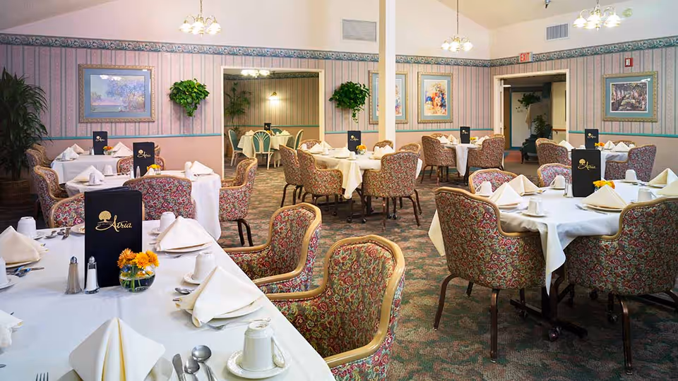A dining room with multiple round tables covered with white tablecloths, each set with folded napkins, cups, and menus labeled 'Atria'. The chairs have a floral pattern upholstery. The room has patterned wallpaper, framed artwork on the walls, and hanging light fixtures. There are plants placed around the room and an exit sign above a doorway.