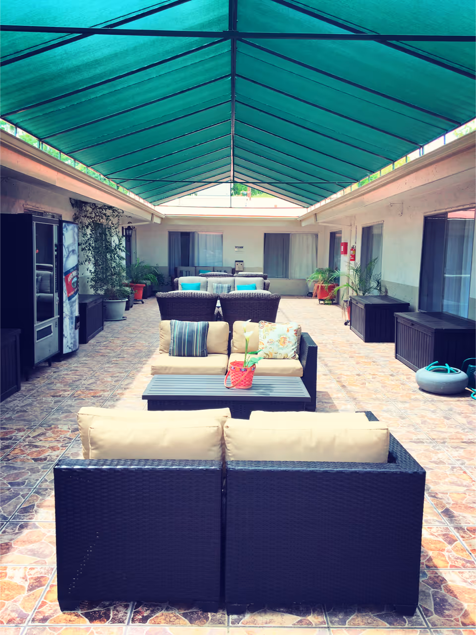 Covered outdoor patio area with wicker seating including sofas and chairs arranged around coffee tables. The floor is tiled, and there are potted plants along the walls. The ceiling is covered with a green canopy providing shade.