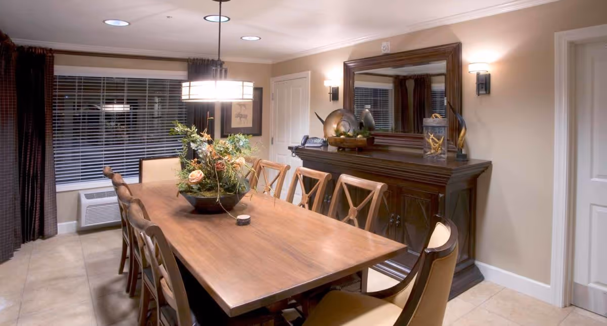 A well-lit dining room with a long wooden table surrounded by eight chairs. A decorative floral centerpiece is placed on the table. The room features beige walls, a large mirror above a dark wooden sideboard, and two wall sconces. A window with blinds and dark curtains is visible on the left side.