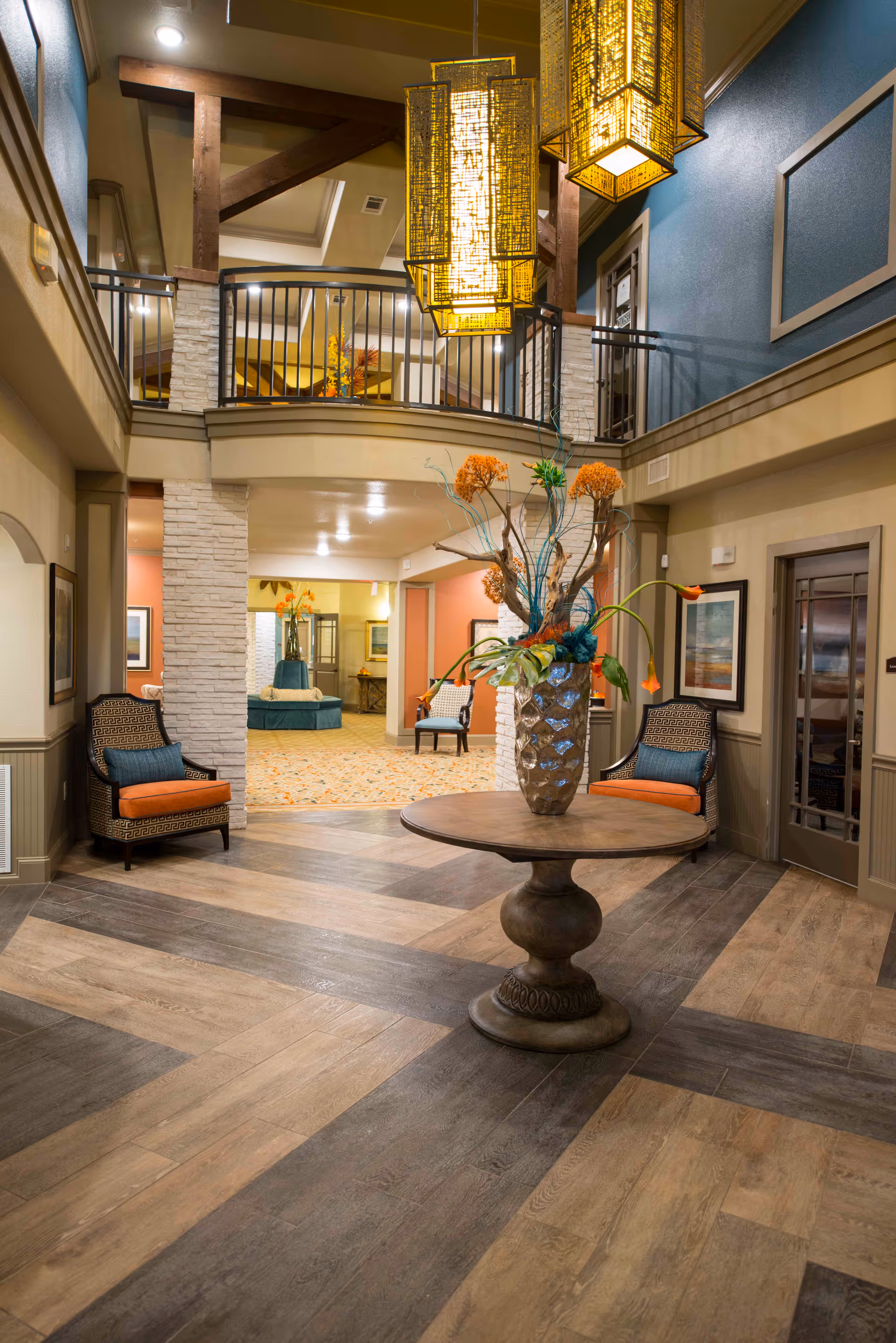 Interior view of a senior living facility lobby with a round wooden table in the center holding a large decorative vase with orange and green floral arrangement. The space features wood-patterned flooring, two cushioned chairs with orange and blue pillows on either side, a second-floor balcony with black railing, and warm hanging pendant lights. The walls are painted in neutral and blue tones with framed artwork.