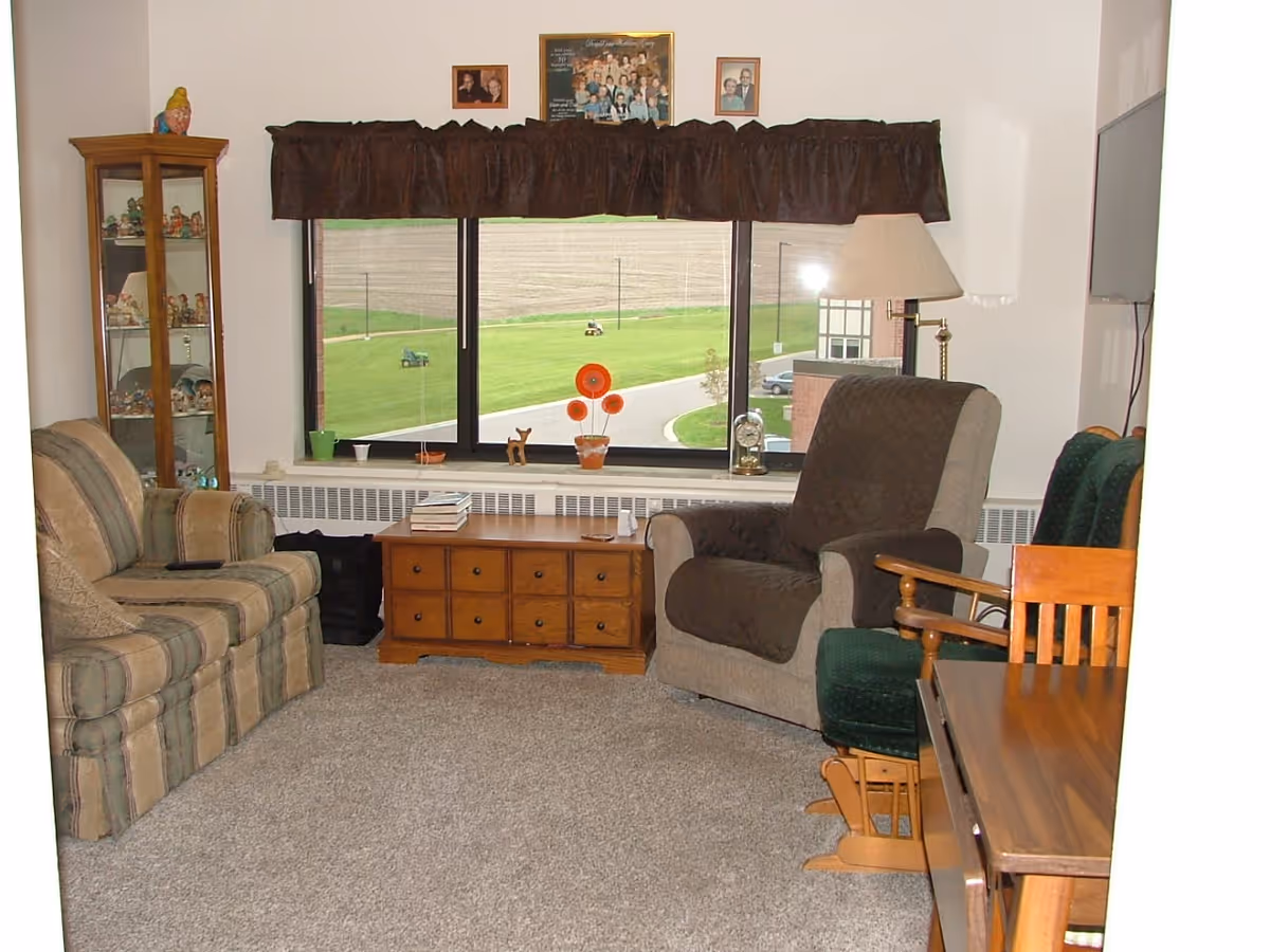 A cozy living room in a retirement community featuring a striped sofa on the left, a cushioned armchair with a brown cover on the right, a wooden coffee table with drawers in front of a large window with a brown valance. The window overlooks a green lawn and a road. A tall glass display cabinet with figurines is on the left, and a floor lamp is next to the armchair. A wooden dining table and chairs are partially visible on the right side.