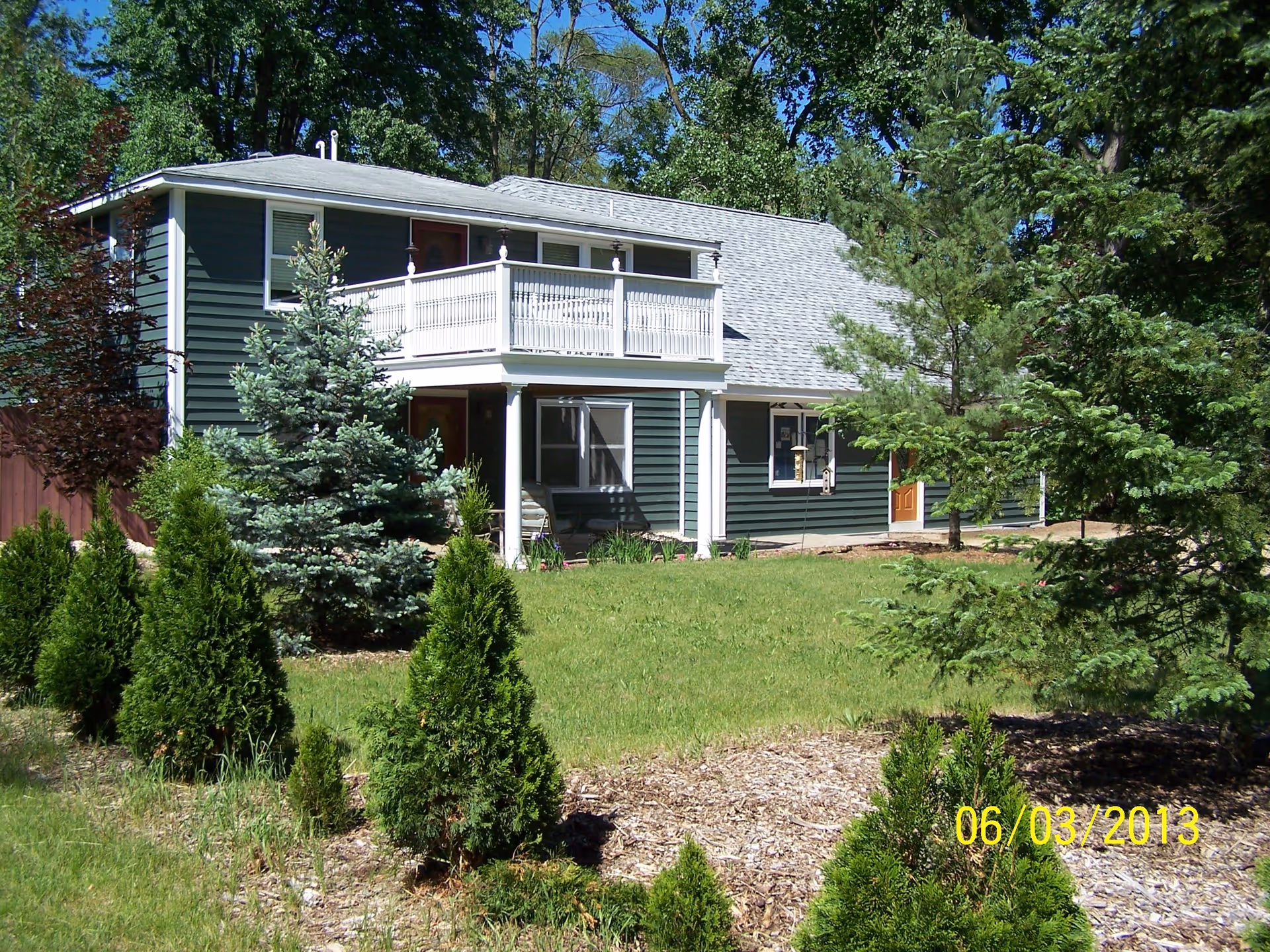 Exterior view of a two-story green house with white trim surrounded by trees and shrubs under a clear blue sky.
