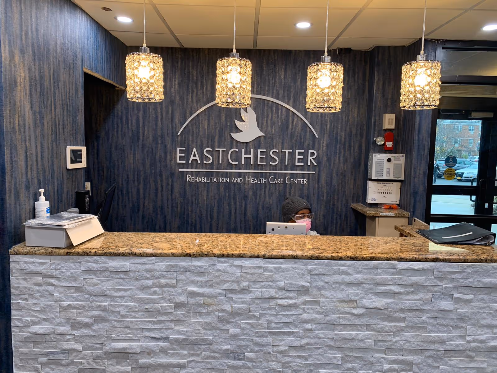 Reception desk area inside Eastchester Rehabilitation and Health Care Center with a granite countertop and white stone facade. Behind the desk is a dark blue textured wall with the facility's name and logo featuring a dove. Four decorative pendant lights hang from the ceiling. A person wearing a pink face mask and a dark hat is seated behind the desk. To the right, there is a glass door leading outside with parked cars visible.