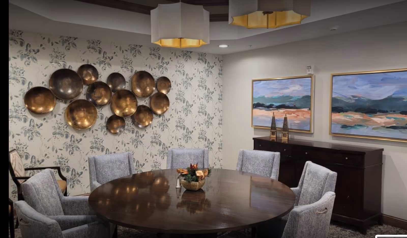 A dining room with a large round wooden table surrounded by six upholstered chairs. The wall behind the table features decorative metallic circular wall art and floral wallpaper. On the adjacent wall, there are two framed landscape paintings above a dark wooden sideboard with decorative items on top. Two modern pendant lights hang from the ceiling.