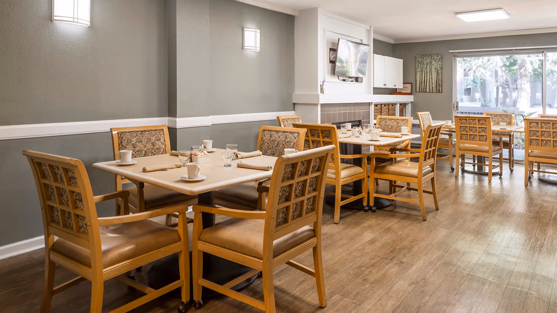 A dining room with several wooden tables and chairs arranged neatly. Each table is set with cups, glasses, and napkins. The room has wooden flooring, light gray walls with white trim, a wall-mounted TV above a fireplace, and large windows letting in natural light.