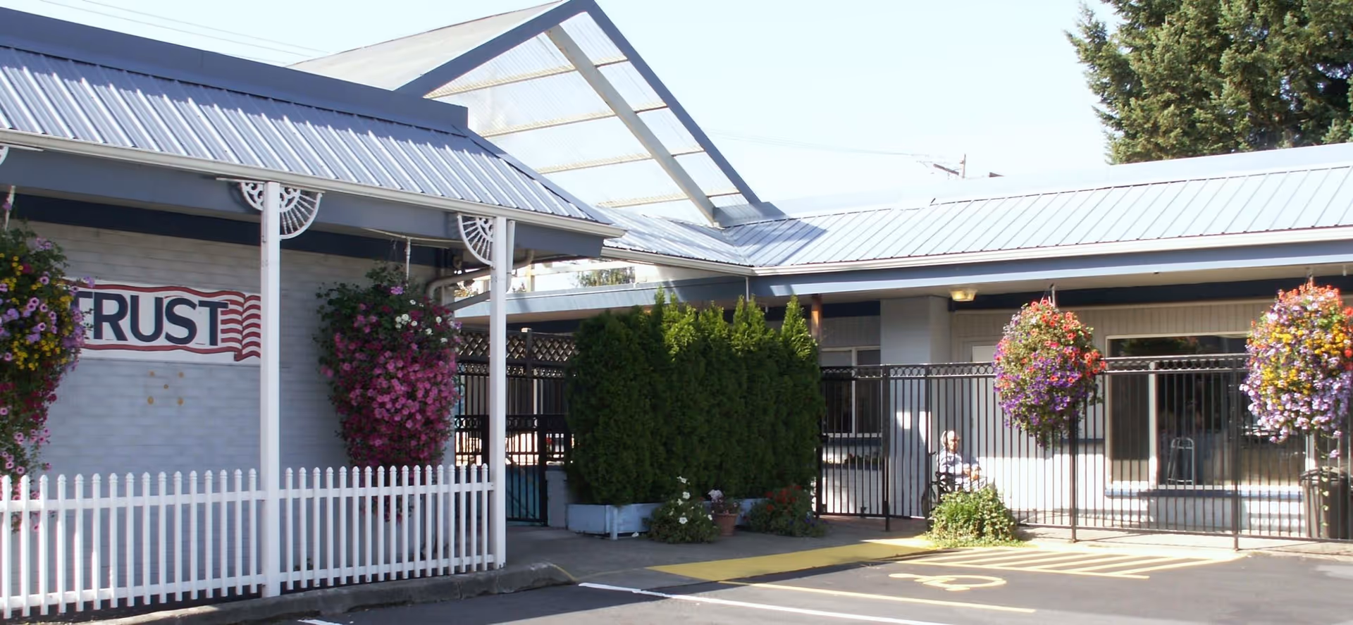 Exterior view of a senior living facility with a covered walkway, hanging flower baskets, a white picket fence, and a gated entrance. A person is sitting near the entrance under the covered area. The building has a metal roof and light-colored walls.