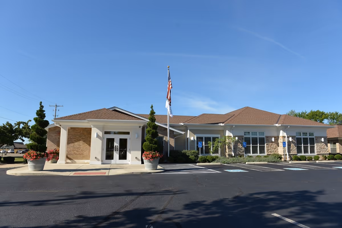 Exterior front view of The Laurels of Hilliard senior living facility building with a clear blue sky, two large potted plants with flowers flanking the entrance, an American flag and another flag on a flagpole, and several handicap parking spaces in front.
