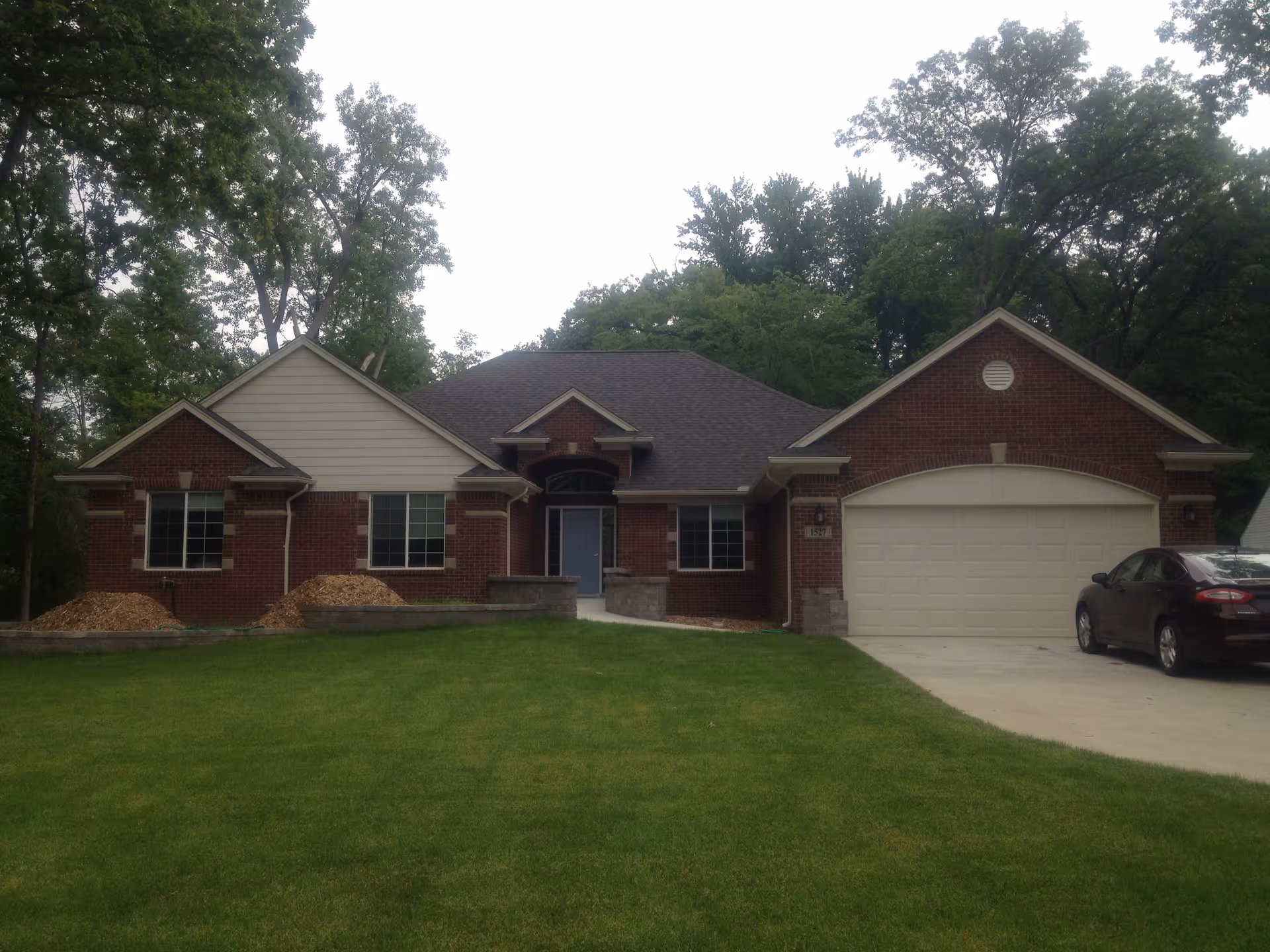 Front exterior view of a single-story brick house with a dark roof, three windows, a blue front door, a two-car garage, a driveway with a parked car, and a well-maintained green lawn surrounded by trees.