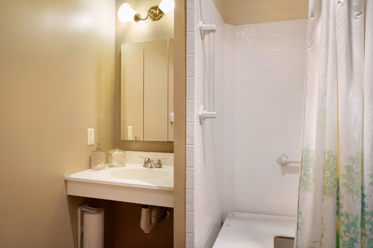 A bathroom with a white sink and countertop, a wall-mounted mirror above the sink, a soap dispenser and a decorative container on the counter. To the right is a white tiled shower area with a grab bar and a floral-patterned shower curtain.