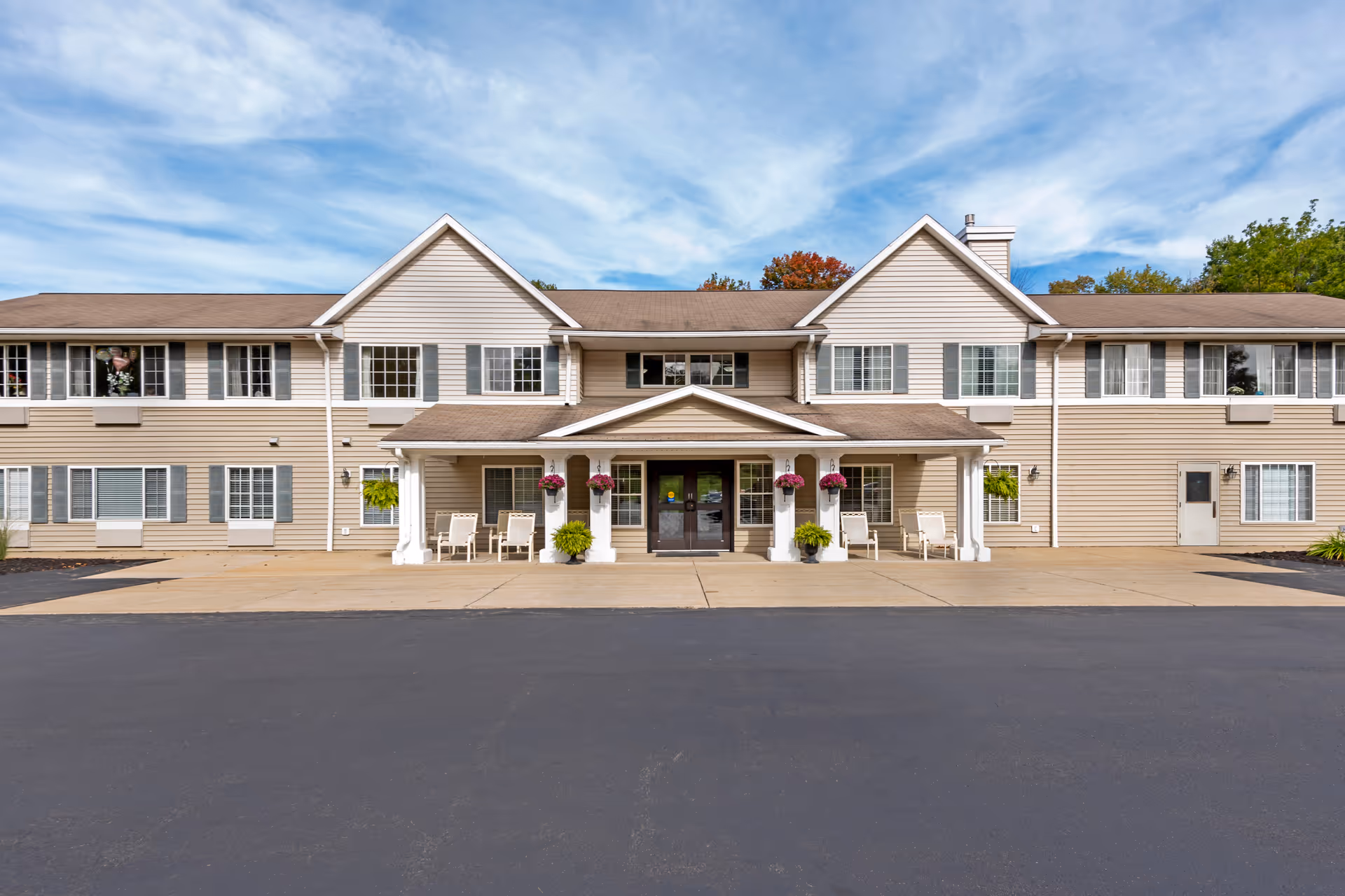 Front exterior view of a two-story senior living facility building with beige siding, multiple windows with shutters, a covered entrance with white columns, hanging flower baskets, and chairs on the porch. The sky is partly cloudy.