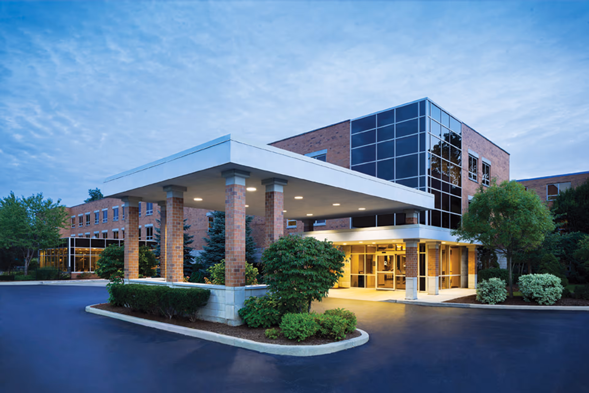 Exterior view of a modern senior living facility building named Glenview Terrace during dusk, featuring a covered entrance with brick pillars, large glass windows, well-maintained landscaping with bushes and trees, and a paved driveway.
