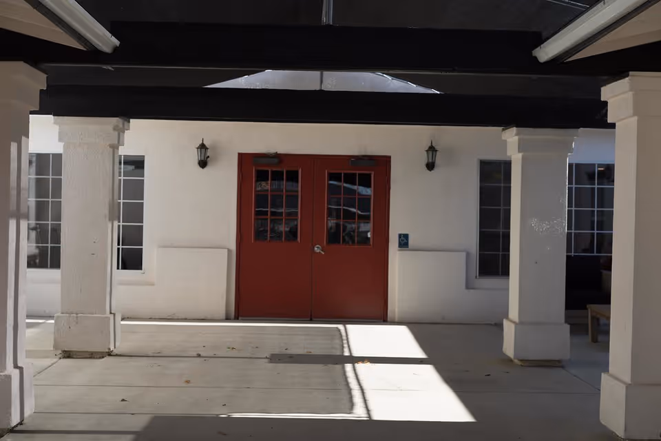 Covered entrance area with white pillars and a pair of red double doors with glass panes. There are two wall-mounted lantern-style lights on either side of the doors and windows on both sides of the entrance. A wheelchair accessibility sign is visible to the right of the doors.