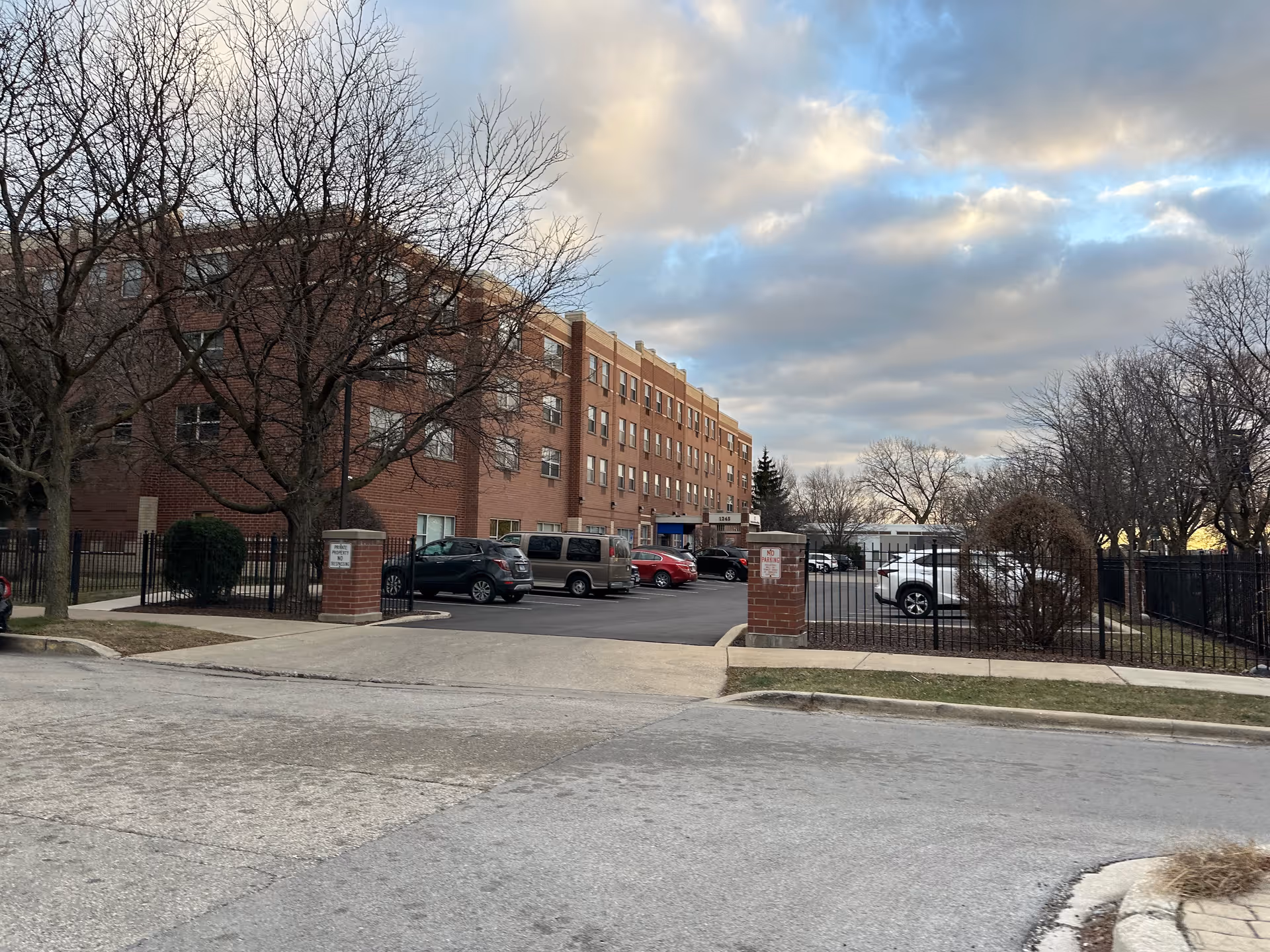 Exterior view of a multi-story brick senior living facility with a parking lot in front. Several cars are parked, and leafless trees surround the area under a cloudy sky.