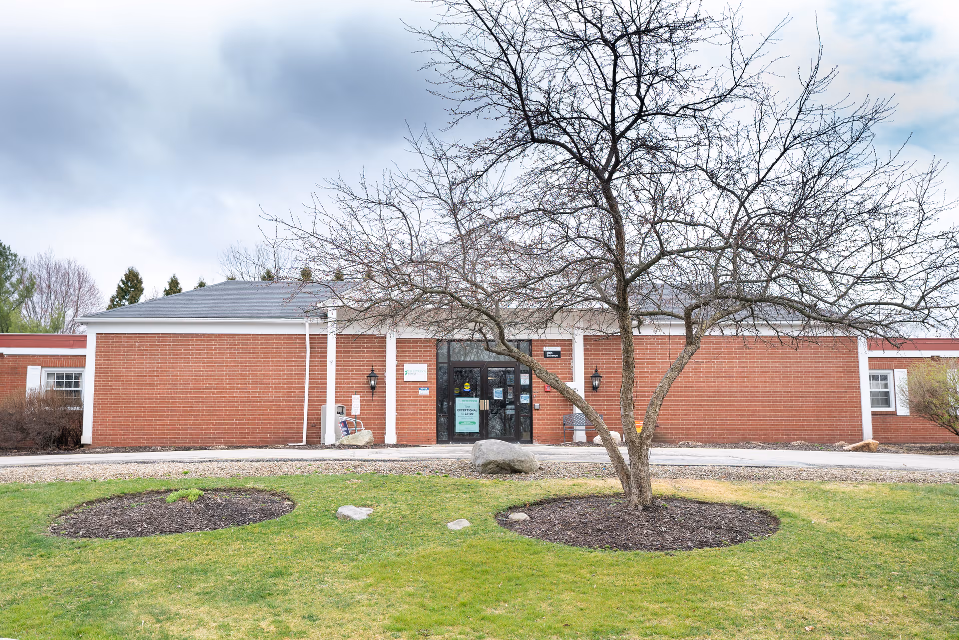 Exterior view of a single-story brick building with a gray roof, a leafless tree in front, and a green lawn with mulched circular planting areas. The entrance has glass doors with signs posted on them.