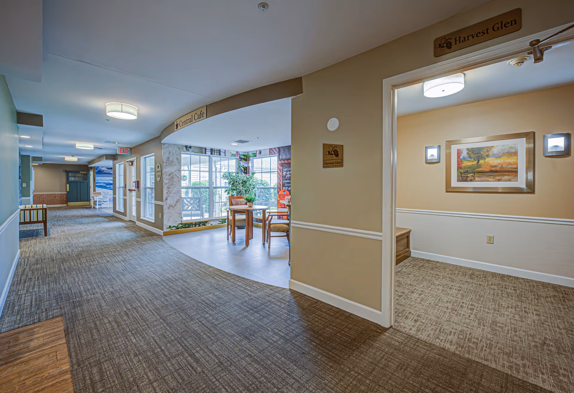 Interior hallway of Arden Courts - ProMedica Memory Care Community with carpeted floor, beige walls, and ceiling lights. To the right is an open room labeled 'Harvest Glen' with a framed landscape painting and two wall sconces. Ahead is a seating area near large windows with a table and chairs under a sign that reads 'Central Cafe'.