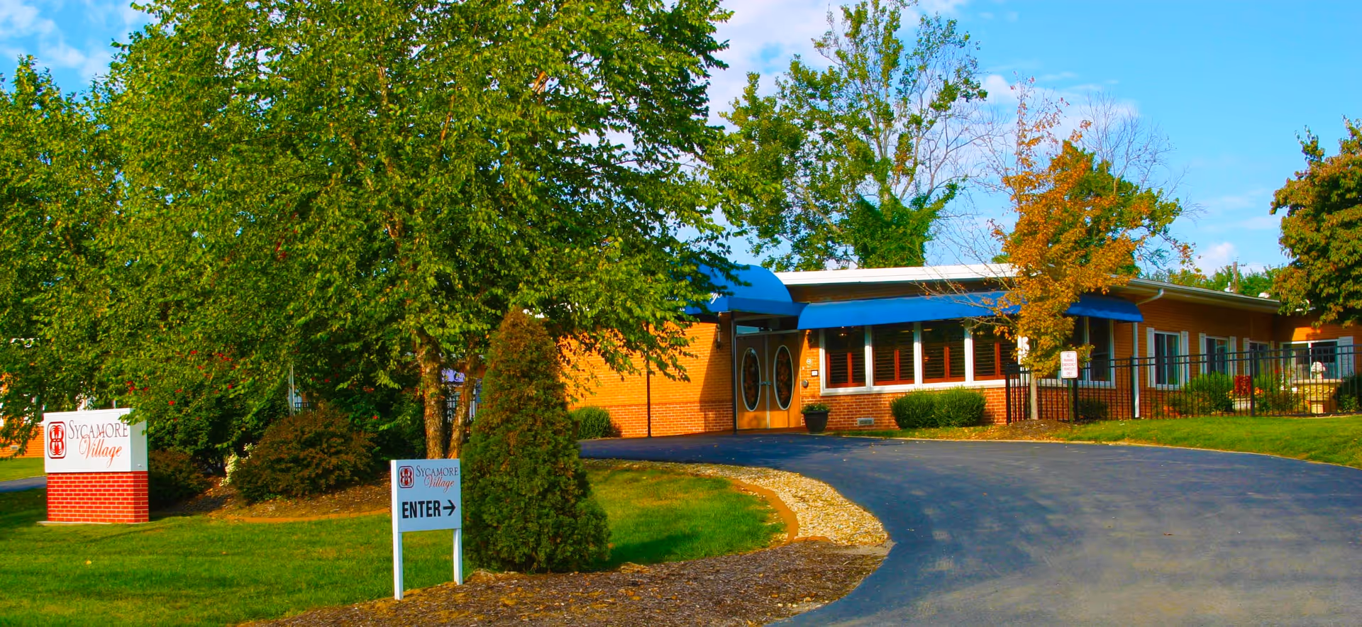 Exterior view of Sycamore Village Assisted Living facility showing a single-story brick building with blue awnings over the entrance and windows, surrounded by green trees and a curved driveway leading to the entrance.