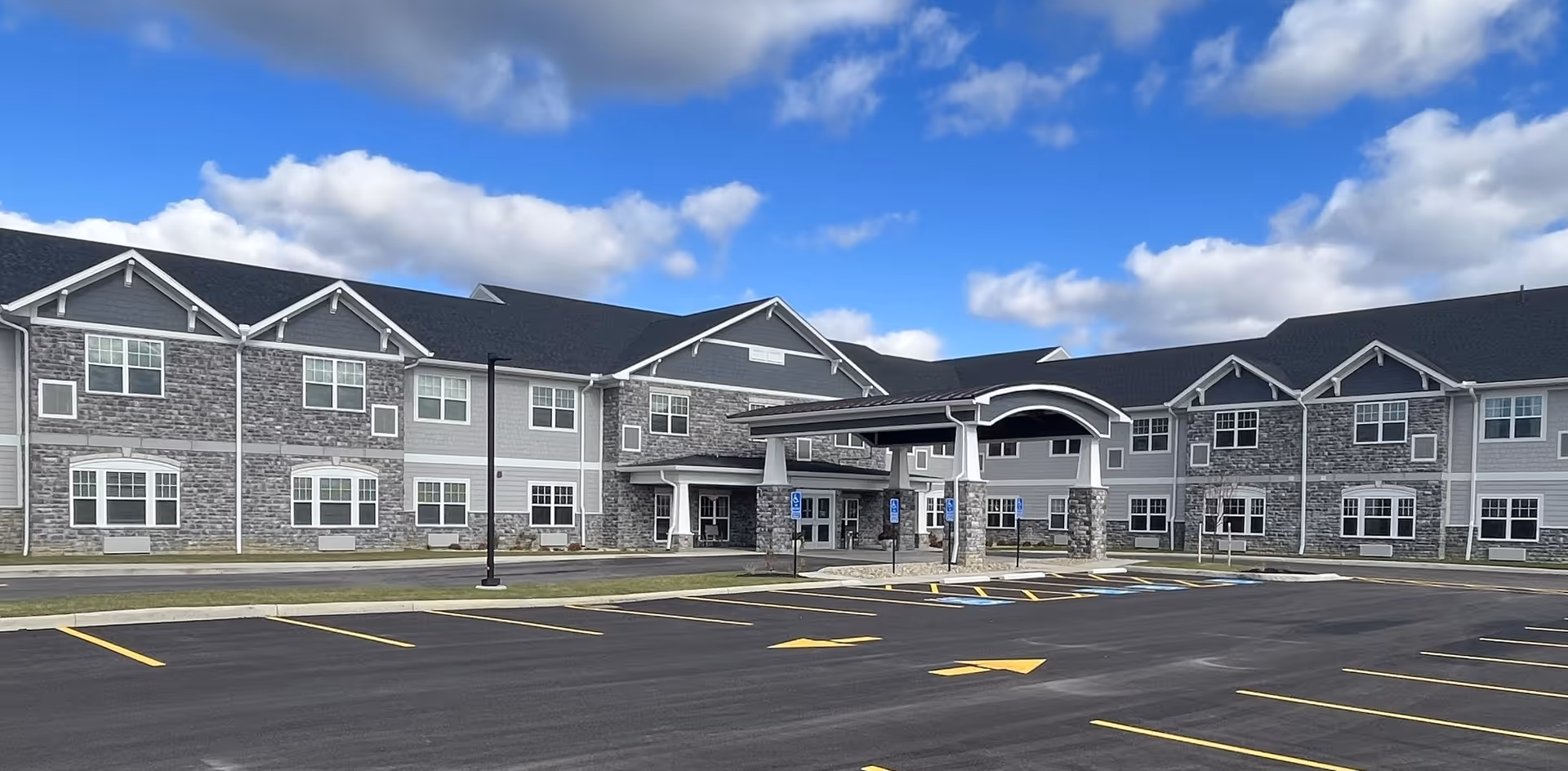 Exterior view of a two-story senior living facility building with gray stone and siding facade, multiple windows, a covered entrance, and an empty parking lot with marked spaces and handicap spots under a partly cloudy blue sky.
