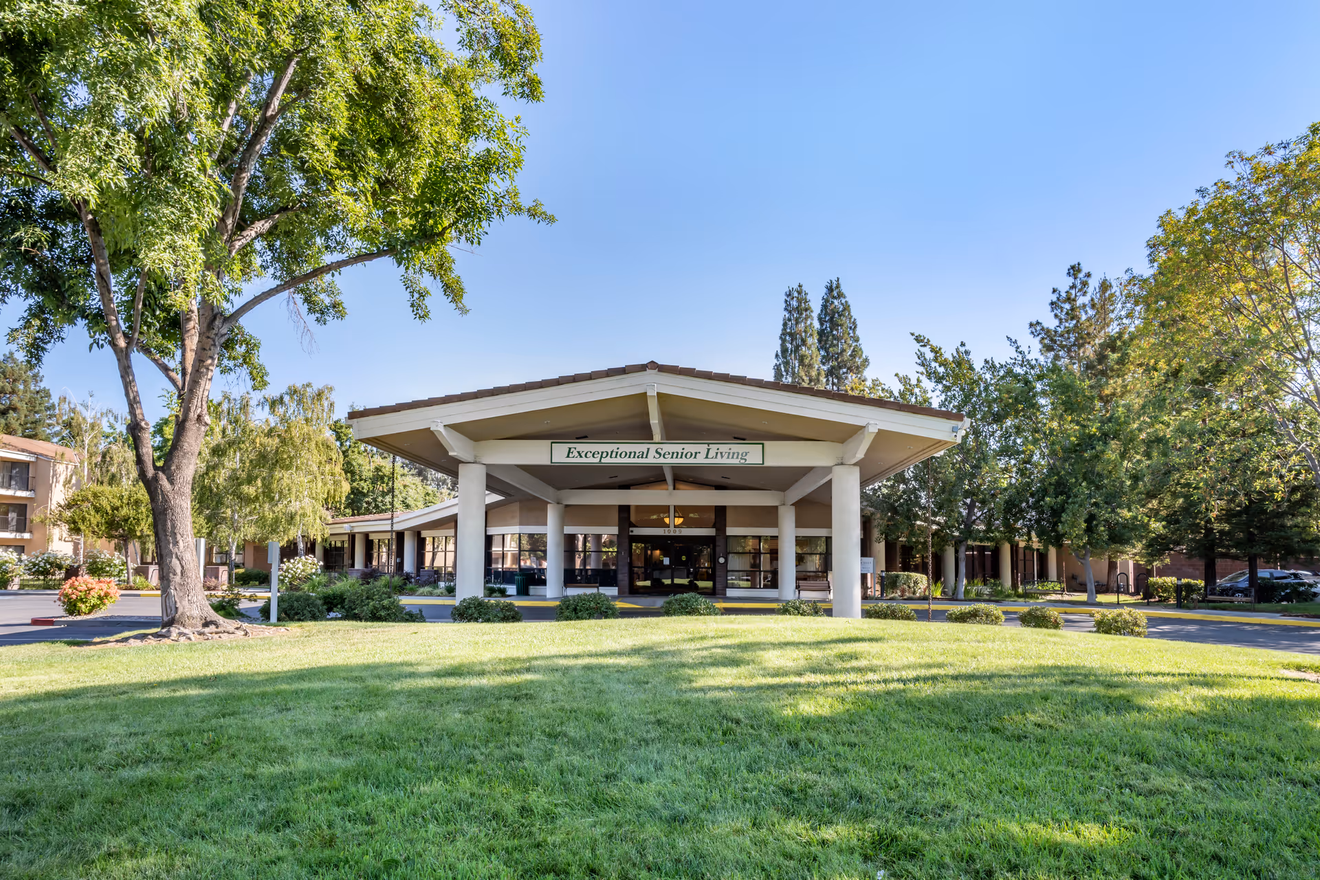 Front exterior view of a senior living facility with a large covered entrance supported by white columns. The building is surrounded by green grass, trees, and shrubs under a clear blue sky. A sign above the entrance reads 'Exceptional Senior Living.'