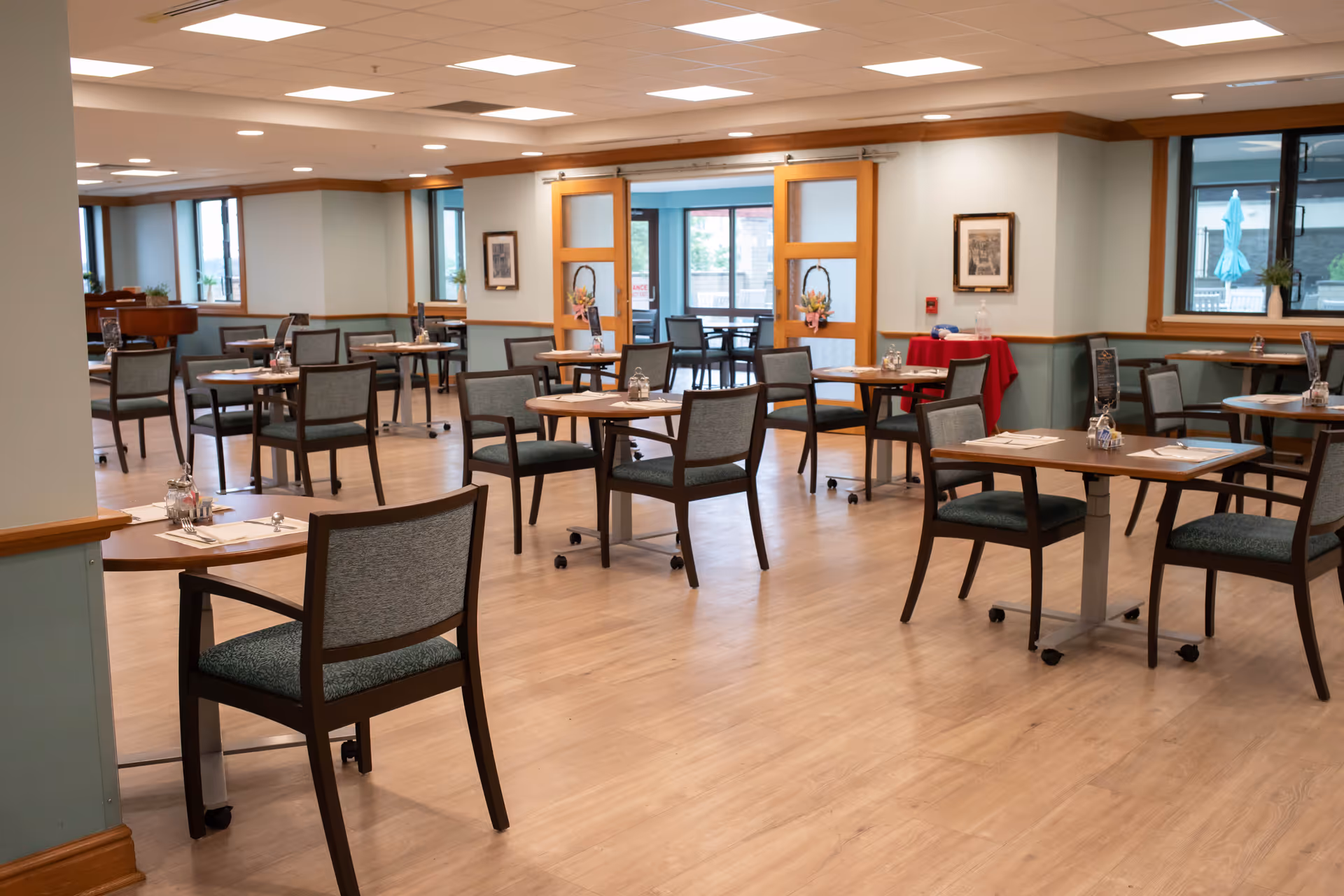 A spacious dining room with multiple wooden tables and chairs arranged neatly. The room has light-colored wooden flooring, soft blue walls with wooden trim, and large windows letting in natural light. There are sliding wooden doors with glass panels decorated with floral wreaths, and a red table with a hand sanitizer dispenser near the wall.