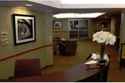 Reception area of a senior living facility with a curved wooden desk in the foreground, a vase with white flowers on the desk, and a seating area with chairs and framed artwork on the walls in the background.