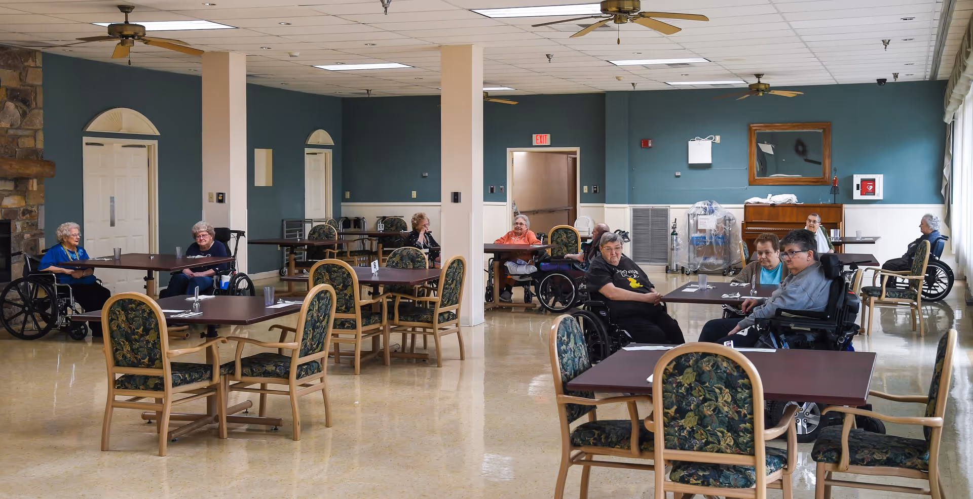 A spacious dining room in a senior living facility with several elderly residents, many in wheelchairs, seated at tables with floral upholstered chairs. The room has a polished floor, ceiling fans, and teal-colored walls with doors and a large window letting in natural light.