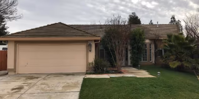 Single-story residential house with a two-car garage, beige exterior walls, and a brown tiled roof. The front yard has a green lawn, some bushes, and small trees. The sky is overcast.