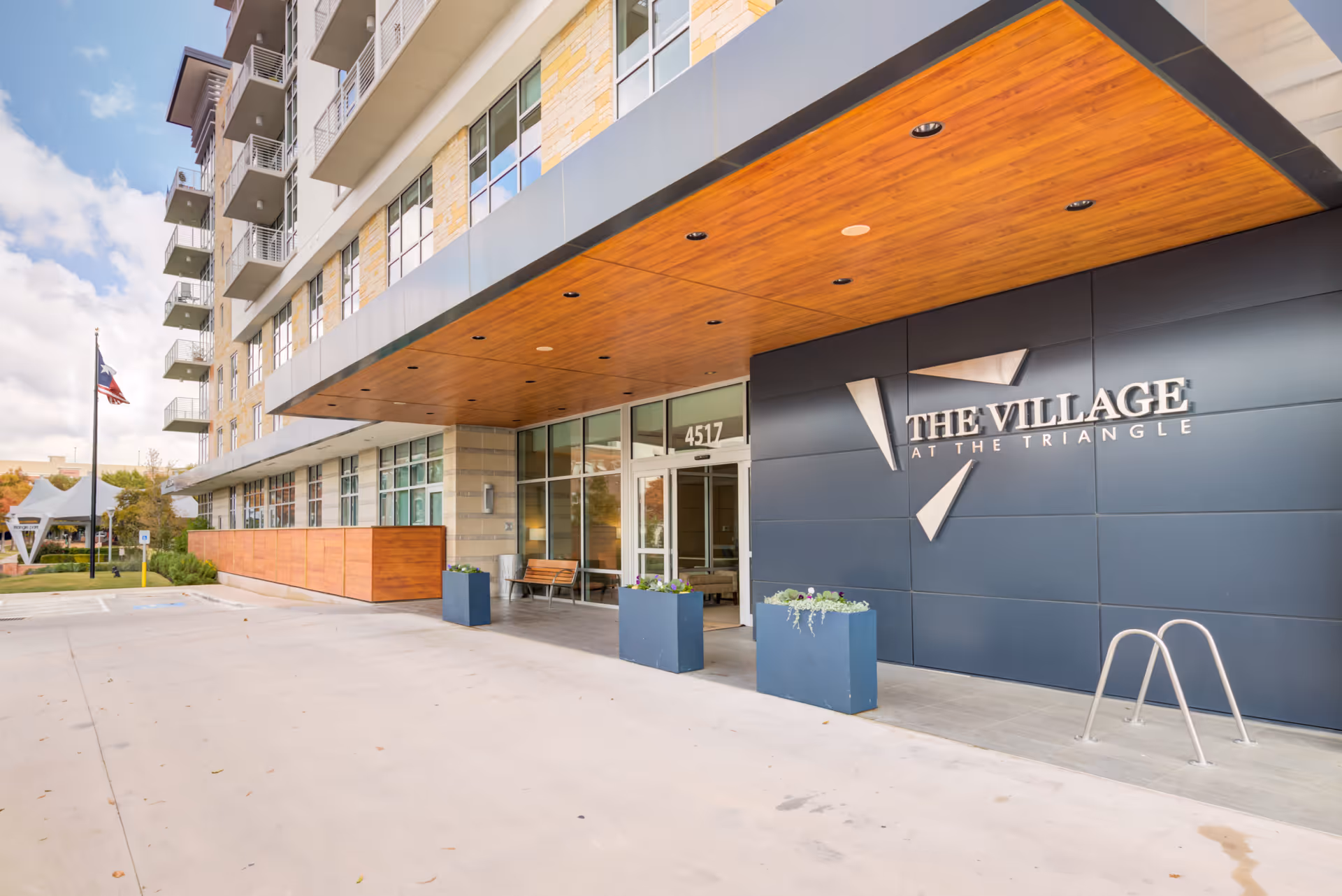 Exterior entrance of The Village at The Triangle senior living facility with a modern building facade, large windows, a wooden ceiling overhang, three blue planters with flowers, a bench, and a Texas state flag on a flagpole in the background.