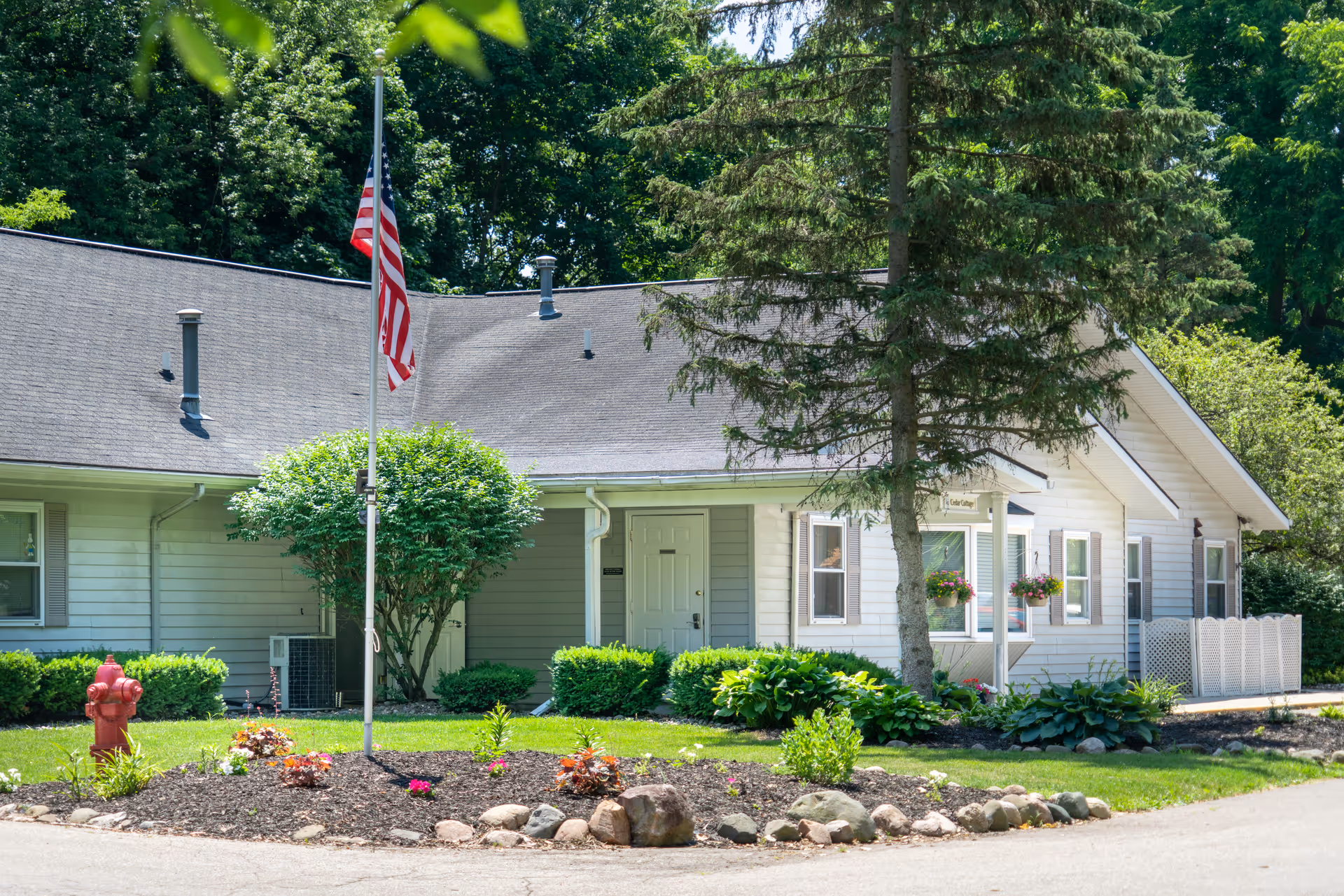 Exterior view of a single-story residential building with light gray siding and a dark gray roof, surrounded by green trees and shrubs. A flagpole with an American flag stands in front of the building, along with a landscaped area featuring rocks, plants, and a red fire hydrant.