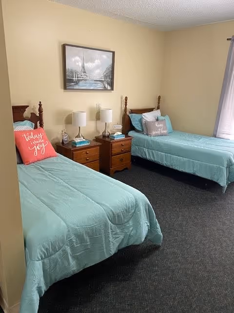 A bedroom with two twin beds featuring light blue bedding and decorative pillows. Between the beds are two wooden nightstands with lamps, a clock, and a tissue box. A framed picture of a sailboat on water hangs on the beige wall above the nightstands. A window with curtains is visible on the right side of the room.