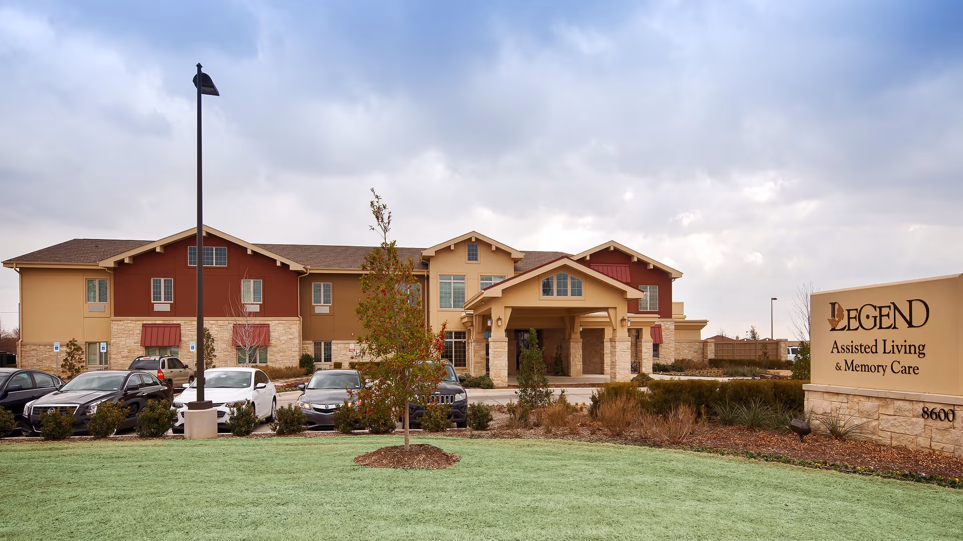 Exterior view of Legend at Fort Worth, an assisted living and memory care facility, showing a two-story building with a covered entrance, parked cars, a lawn with small trees, and a large sign displaying the facility's name and services.