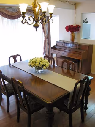 A dining room with a wooden table and six chairs, a white table runner, and a centerpiece of white flowers. A chandelier with six lights hangs above the table. In the background, there is a wooden piano with a vase of red flowers on top, and a window with sheer curtains and drapes.
