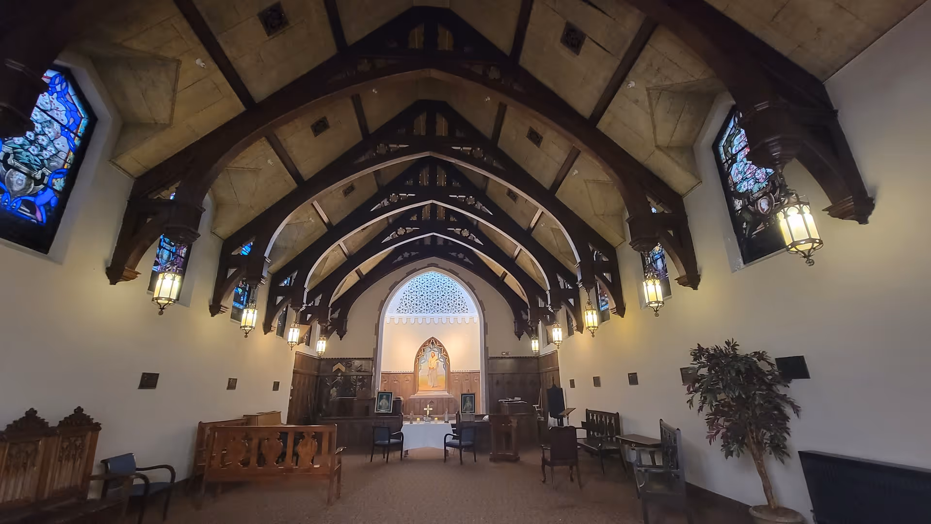 Interior view of a chapel with wooden arched ceiling beams, stained glass windows on both sides, hanging lantern-style lights, wooden pews, and a religious altar at the far end featuring a painting and a cross.