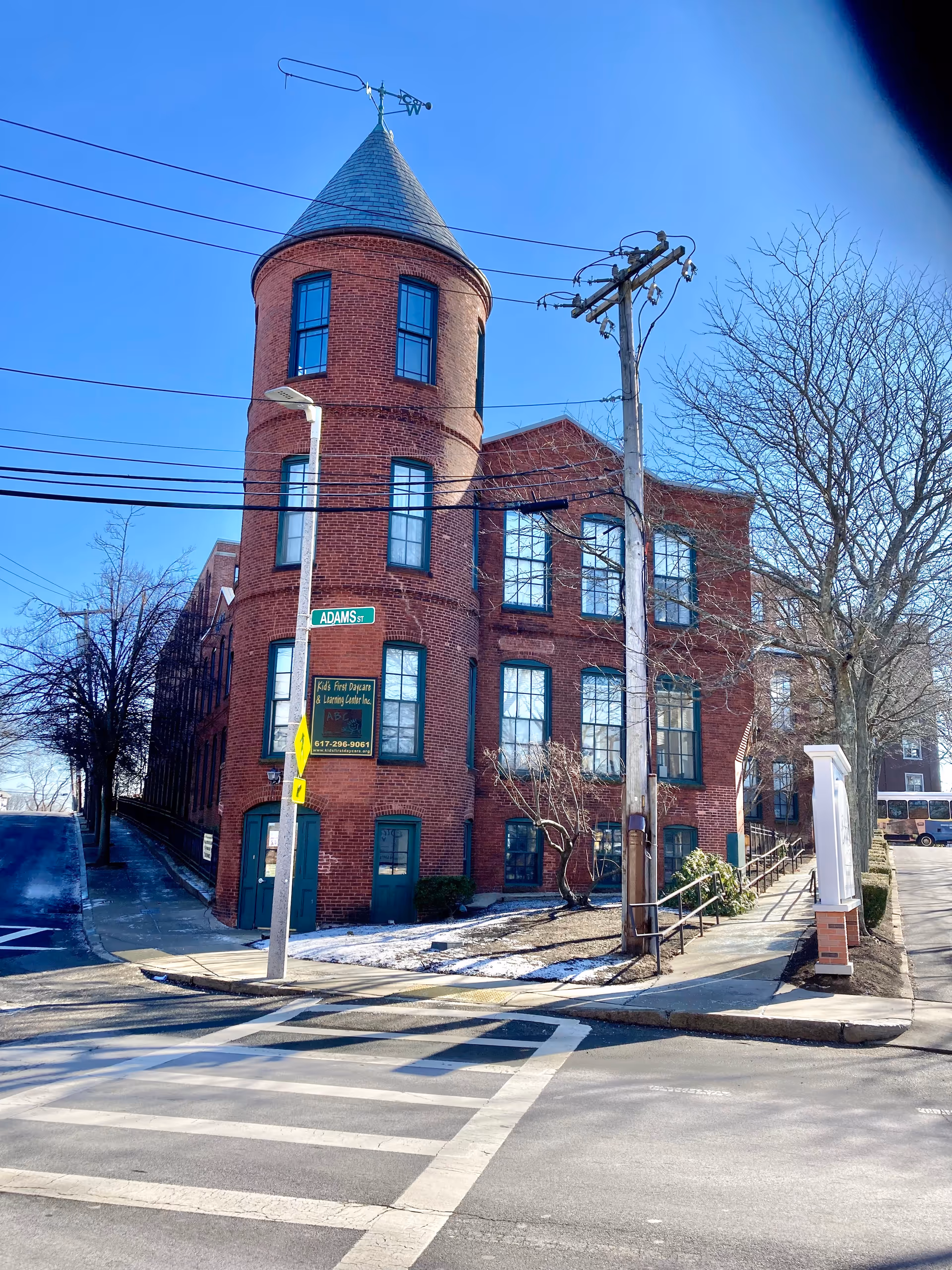 A red brick building with a rounded turret and multiple windows on a clear day. The building is located at the corner of Adams Street, with a street sign and a lamppost visible. There is a sidewalk and a crosswalk in the foreground, and some leafless trees and a utility pole nearby.