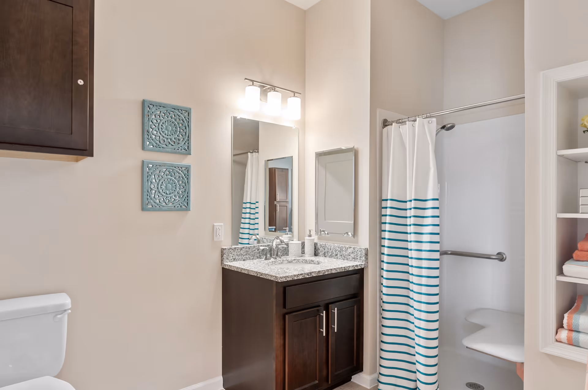 A clean and modern bathroom featuring a white toilet, a dark wood vanity with a granite countertop and a sink, a large mirror with three light fixtures above it, a shower with a white and teal striped curtain, and built-in shelves holding folded towels and decorative items.