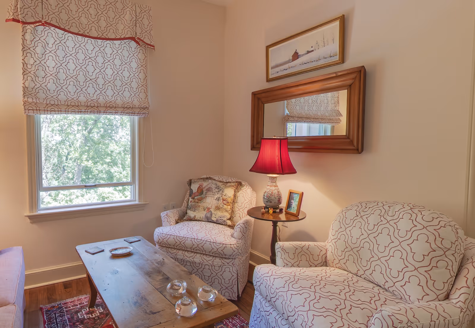 A cozy living room corner with two patterned armchairs and a wooden coffee table. A small round side table between the chairs holds a decorative lamp with a red shade and a framed photo. A large window with patterned curtains lets in natural light, and a framed painting hangs above a wooden-framed mirror on the wall.