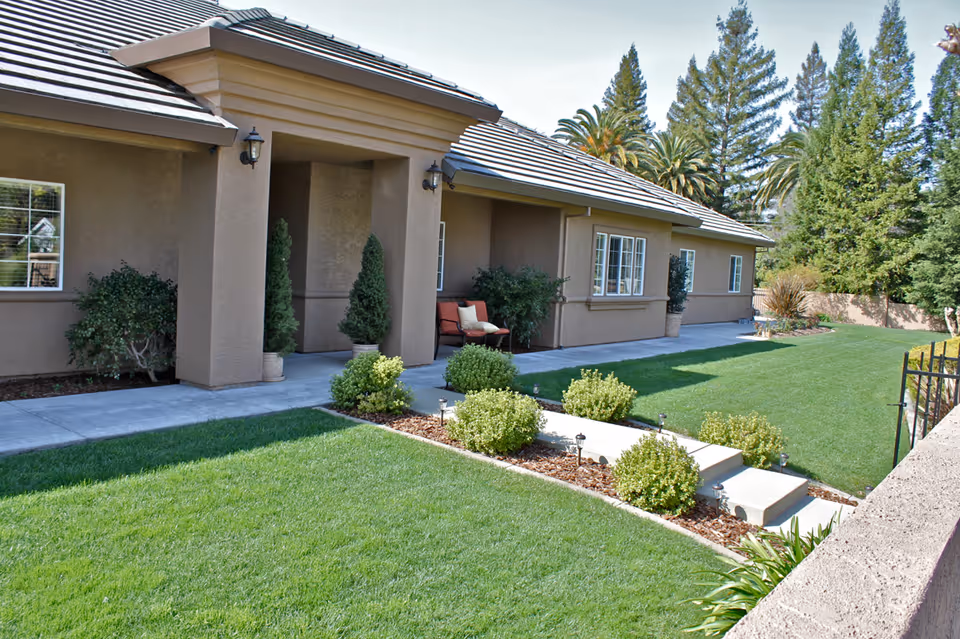 Exterior view of a single-story building with a beige stucco finish and a tiled roof. The building has a covered porch area with two potted plants and a bench with cushions. The surrounding area features a well-maintained green lawn, small bushes, and trees in the background under a clear sky.