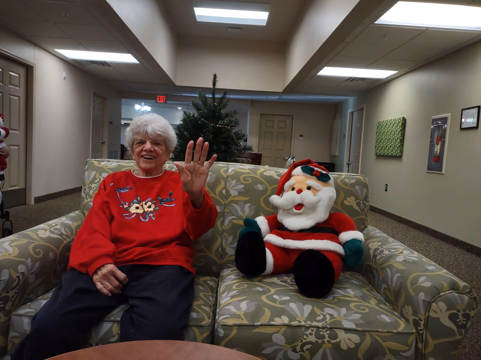 An elderly woman wearing a red sweater with floral embroidery is sitting on a patterned couch next to a large stuffed Santa Claus doll. She is smiling and waving with her right hand. Behind her, there is a Christmas tree and a hallway with doors and framed pictures on the walls.