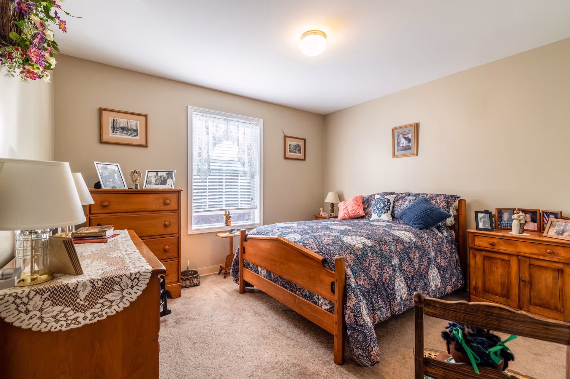 A cozy bedroom with a wooden bed frame and a patterned blue and pink bedspread. The room has beige walls and carpeted floor. There are two wooden dressers, one with a lace runner and a lamp, and the other with framed photos and a small statue. A window with white blinds and lace curtains lets in natural light. Several framed pictures hang on the walls, and a wooden chair with a doll is partially visible in the foreground.