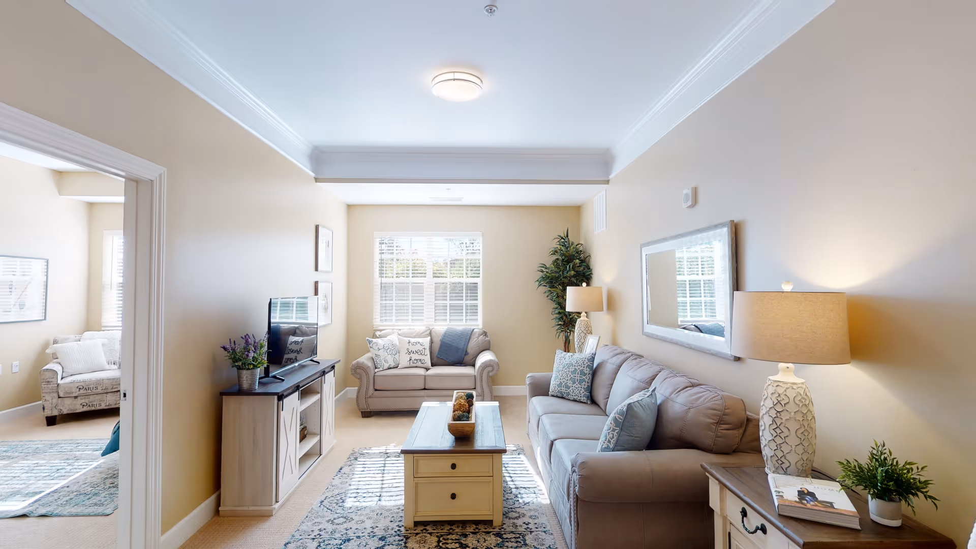 A bright and cozy living room with beige walls and carpet. The room features a gray sofa with decorative pillows, a matching loveseat near a window with white blinds, a wooden coffee table with decorative items, a side table with a lamp and a small plant, and a TV on a wooden stand. A large mirror hangs on the wall above the sofa, and a tall potted plant is placed in the corner near the window. An adjacent room with additional seating is visible through an open doorway.