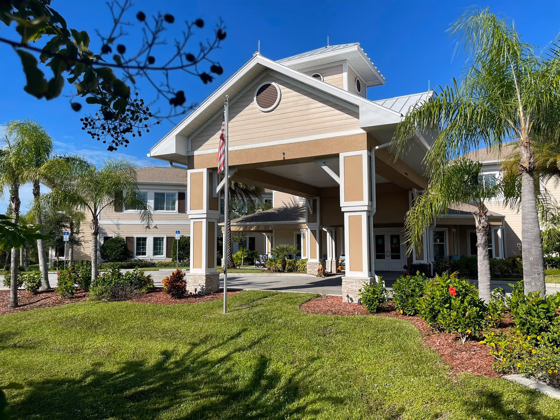 Exterior view of Pelican Landing Assisted Living & Memory Care facility showing the entrance with a covered drop-off area, surrounded by palm trees and landscaped greenery under a clear blue sky.