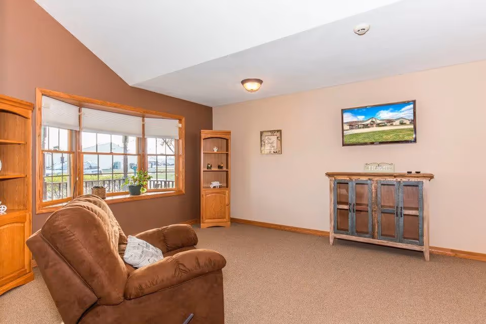 A cozy living room with a brown recliner chair facing a wall-mounted flat-screen TV. The room has beige walls with one accent wall painted brown, a large window with white blinds, and wooden corner shelves with decorative items. There is a wooden cabinet beneath the TV and a carpeted floor.