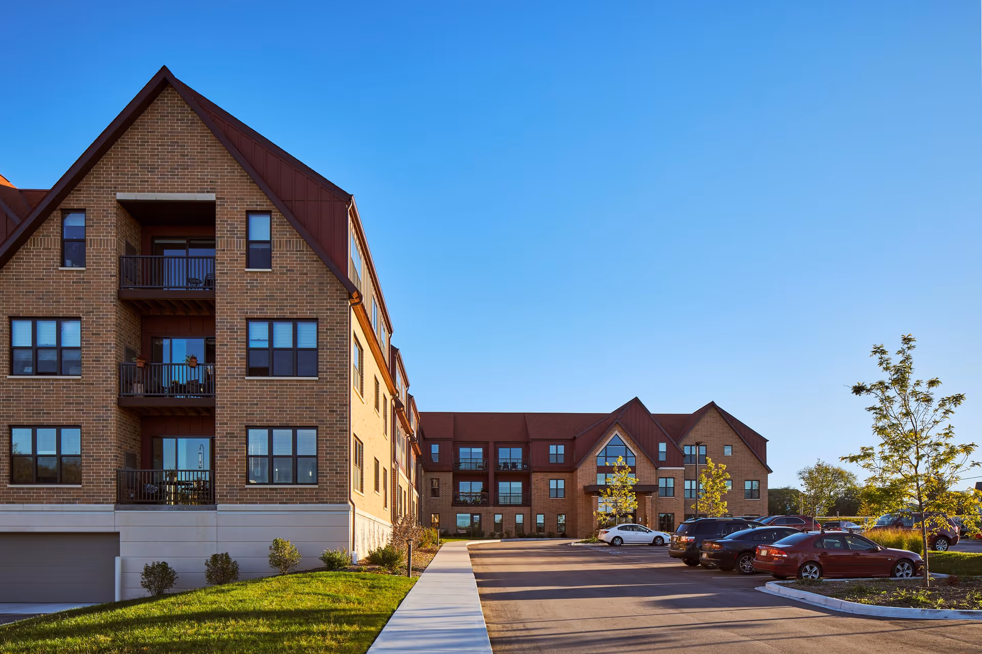 Exterior view of a multi-story brick senior living facility building with balconies, a parking lot with several cars, and a clear blue sky.